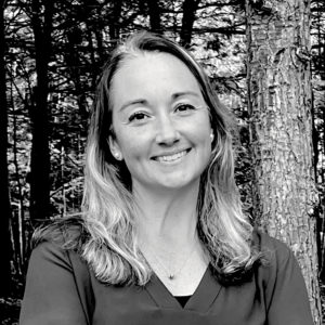 Katie Thompson, a woman with long, light hair, smiles at the camera while standing outdoors in front of trees and a tree trunk. The black and white image captures her joyful expression.