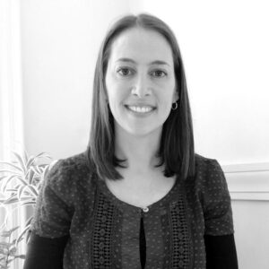 A woman with straight, shoulder-length hair smiles at the camera. She is wearing a short-sleeved, patterned blouse and small hoop earrings. A plant and a light-colored wall are in the background. The black-and-white photo was taken at Great Schools Partnership.