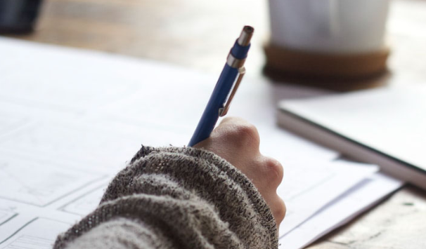 A person in a gray sweater writes with a blue pen on white paper at a desk, reflecting on the assessment continuum, with a blurred notebook and cup in the background.