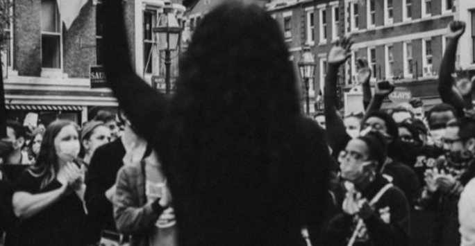 Black and white photo of a person with raised arm facing a crowd on a city street; people in the crowd are clapping, cheering, and some—especially Black youth—are wearing face masks, creating an uplifting scene.
