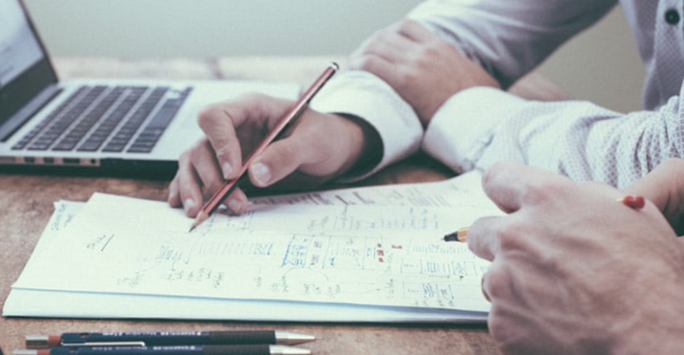 Two people sit at a desk, reviewing handwritten notes and diagrams on paper. One person holds a pencil while the other points with a pen—collaborating on Educating Beyond the Classroom. An open laptop and colored pencils are also on the table.