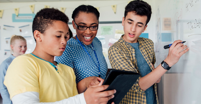 A teacher and two students work together at a whiteboard; one student writes with a marker while the other shows a tablet to the teacher, who is smiling—an inspiring scene of teaching with grace. Another student is blurred in the background.