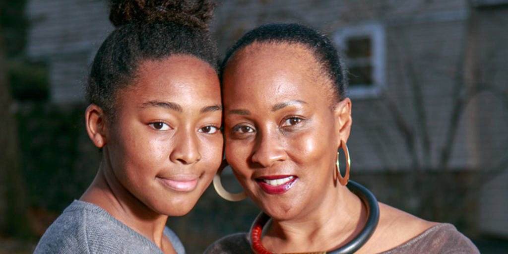 A smiling woman and a teenage girl stand close together outdoors, both looking at the camera, united in their commitment to anti-racism. The woman wears hoop earrings and a necklace; the girl has her hair in a bun. A house is blurred in the background.