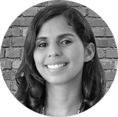 A woman with long, dark hair smiles at the camera in front of a brick wall background. The photo, taken in February 2021, is black and white and shown in a circular frame.