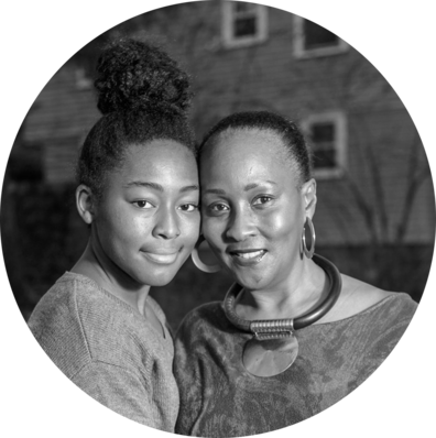 A black and white photo of two women, one younger and one older, standing close together and smiling at the camera. They appear to be outdoors, with a house in the background.