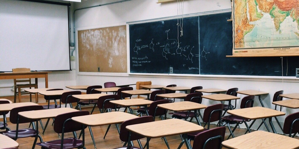 Empty classroom with rows of wooden desks and chairs, a large chalkboard with drawings and notes, a pull-down world map, projector screen, and a teacher’s desk at the front—ready for student engagement or hybrid learning sessions.