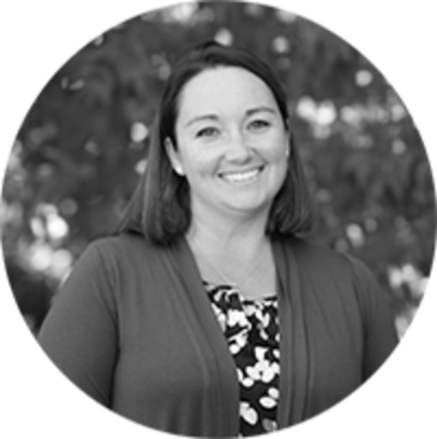 Black and white photo of a woman smiling at the camera, wearing a cardigan over a patterned top, with shoulder-length hair. The background is blurred and appears to be outdoors with trees or foliage.