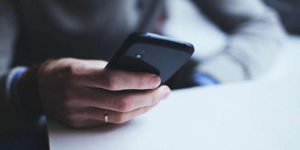 A person wearing a wedding ring holds and uses a smartphone with one hand, sitting at a white table—perhaps exploring advocacy or making your voice heard beyond signing a petition. The background is blurred.