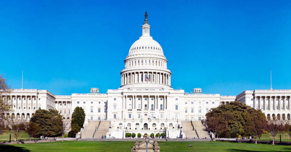 The United States Capitol building under a clear blue sky, home to the Senate Appropriations Subcommittee on Labor Health and Human Services, stands with its iconic white dome and columns, surrounded by green lawns and trees.