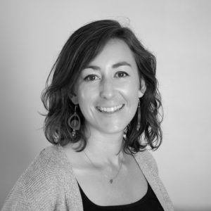 Black and white portrait of Emily Gilmore Hurwitz, a woman with wavy shoulder-length hair, wearing a cardigan, a necklace, and large round earrings, smiling at the camera against a plain background.