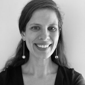Black and white portrait of Gabriela Stiteler, a woman with long dark hair, wearing a v-neck top and dangling earrings. She is smiling and facing the camera against a plain, light background.