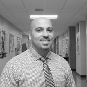 A man with a shaved head, goatee, and checked shirt with a striped tie smiles while standing in a hallway with bulletin boards on the walls. The image is in black and white.