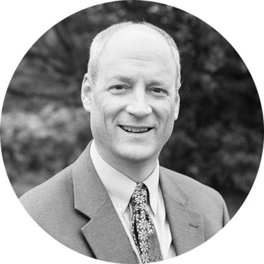 Black and white portrait of a middle-aged man in a suit and patterned tie, smiling at the camera with trees blurred in the background. The image, circularly cropped, was taken in November 2021.