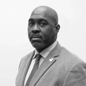A serious-looking man with a bald head and a short beard, wearing a suit, dress shirt, and tie, poses against a plain light background. The image is in black and white.