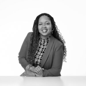 Danielle Pierre, a woman with long curly hair, wearing a patterned blouse and blazer, smiles and leans forward with her hands resting on a white surface. The photo is black and white with a plain background.