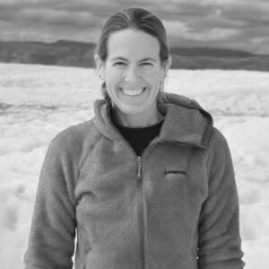 Erica Wallstrom, with long hair pulled back, smiles at the camera in a fleece jacket. Snow or ice fields and distant mountains create a striking black and white backdrop.