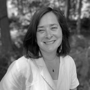 Jeanie Phillips, with shoulder-length dark hair, wearing a light-colored blouse and a necklace, smiles at the camera. The background is blurred trees and foliage in an outdoor setting. The image is in black and white.