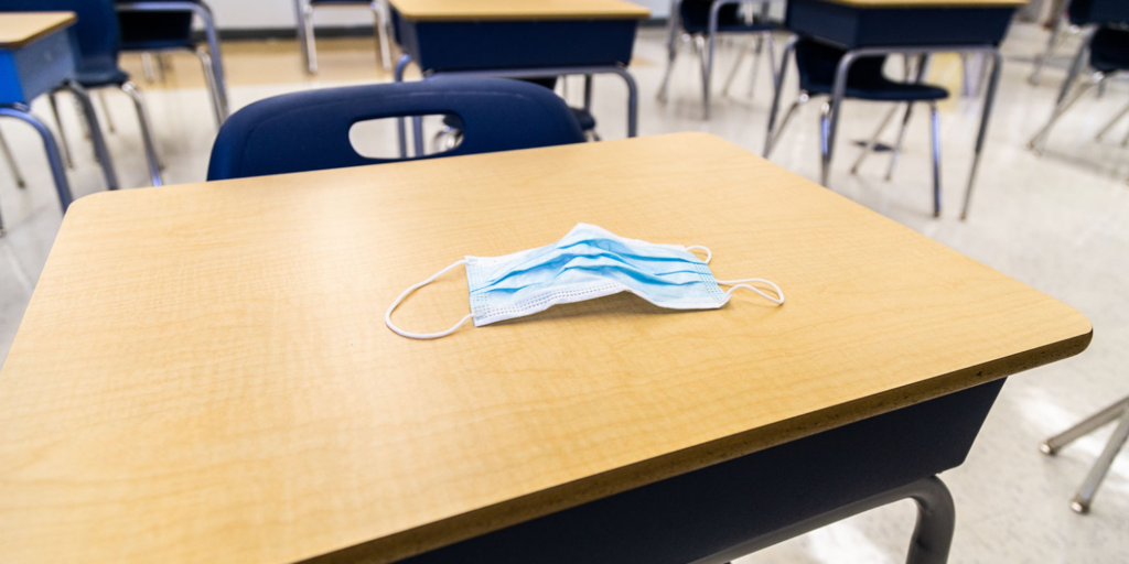 A disposable blue face mask lies on a wooden classroom desk with empty chairs and desks in the background, reflecting how COVID continues to impact learning in unoccupied classrooms.