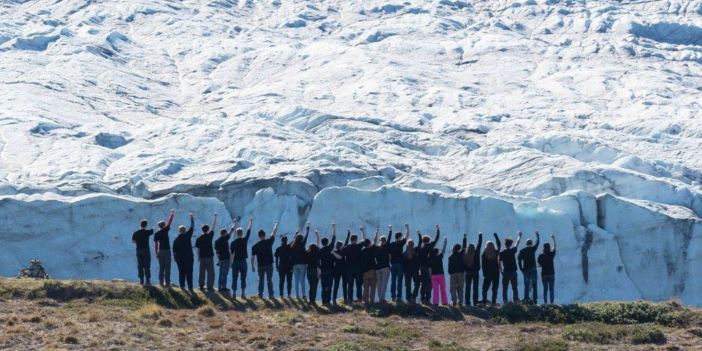 A group of people stand on grassy ground facing a large glacier, raising one arm in the air as if filming a dance video. Everyone is dressed in dark clothing except one person in bright pink pants. The glacier dominates the background.