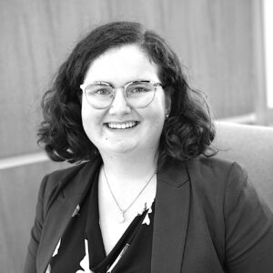 Kayla Girardin, with curly dark hair, wearing glasses, a blazer, and a patterned blouse, is smiling while seated in front of a wooden background.