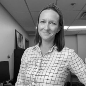 Brean Witmer, a woman with straight, shoulder-length hair and a checked shirt, smiles while standing in an office with a computer, artwork on the wall, and a tiled ceiling. The image is in black and white.