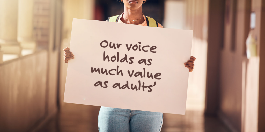 A person holds a sign that says, Our voice holds as much value as adults, standing in a hallway with blurred background—highlighting the importance of students being heard by teachers and school leaders.