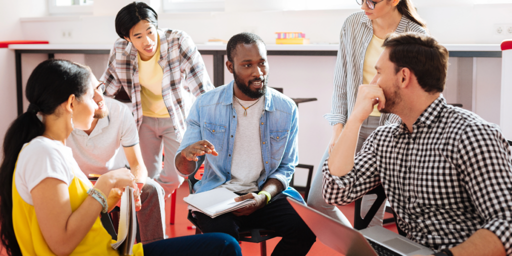 A group of five people are sitting and standing in a bright room, having a discussion. One person in the center speaks while holding a notebook, bringing clarity to the conversation as the others listen attentively.