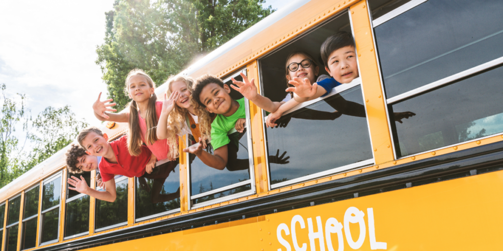 A group of smiling children lean out of the windows of a yellow school bus, waving cheerfully on a sunny day. Their happy attendance highlights the importance of equity in access to education. Trees are visible in the background.