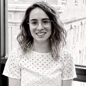 A woman with wavy, shoulder-length hair and glasses smiles indoors by a window, with a brick building outside. She is wearing a white, short-sleeved, eyelet-patterned top—an inviting scene reminiscent of Dylan Reynolds' cozy style.