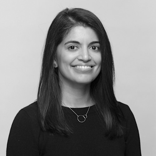 A woman with straight dark hair, wearing a black top and a circular pendant necklace, smiles at the camera against a plain light background. The image is in black and white.