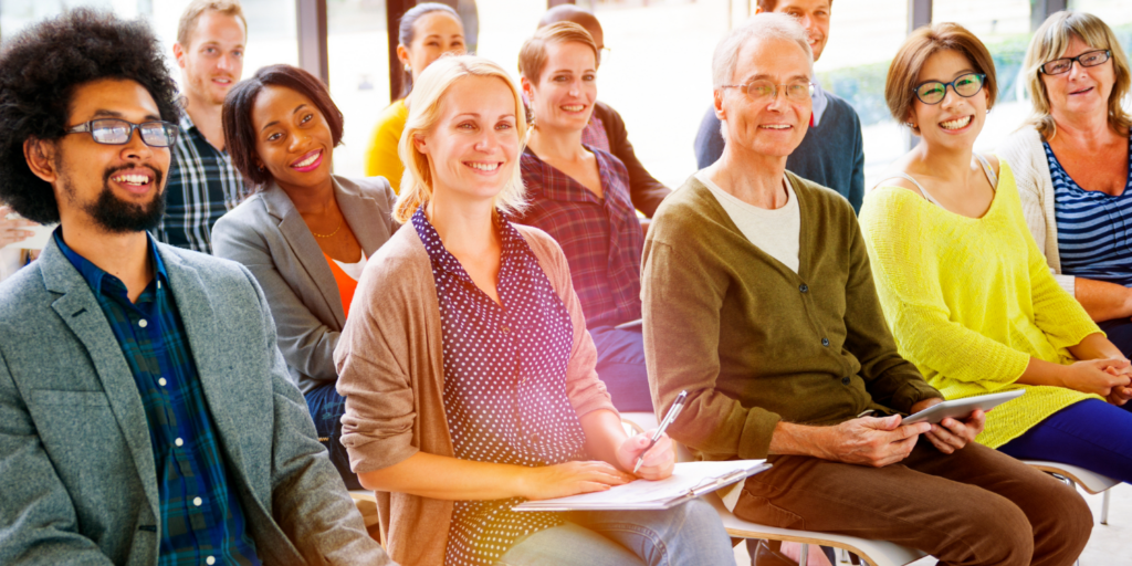 A diverse group of adults sits in rows, smiling and looking toward the front, as if attending a class or seminar. Some hold notebooks or tablets, sunlight streaming in as they engage in a safe and brave learning community.