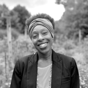 Darthula Mathews, wearing a headwrap, blazer, and necklace, smiles warmly at the camera while standing outdoors in a natural setting with blurred greenery in the background. The photo is in black and white.