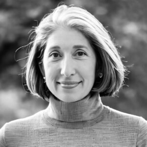 Elisabeth Kanner, a woman with short, light-colored hair wearing a textured turtleneck top, smiles gently at the camera. The blurred outdoor background creates a soft, natural atmosphere.