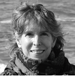Isabella Borrero, with wavy hair, smiles at the camera while standing outdoors in front of a body of water. She is wearing a scarf and a jacket. The black and white image captures her warm expression.