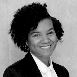 Black and white portrait of Krystal Bravo, a person with short curly hair, wearing hoop earrings, a collared shirt, and a blazer, smiling at the camera against a plain background.