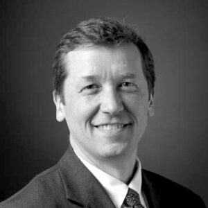 Stephen Bowen, in a suit and tie, smiles at the camera against a plain, dark background in this black and white photo.