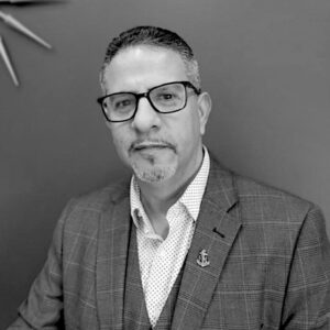 Black and white portrait of Victor Capellan, a man with short hair, glasses, and a goatee, wearing a checkered suit jacket, patterned shirt, and anchor-shaped pin, sitting in front of a plain wall.