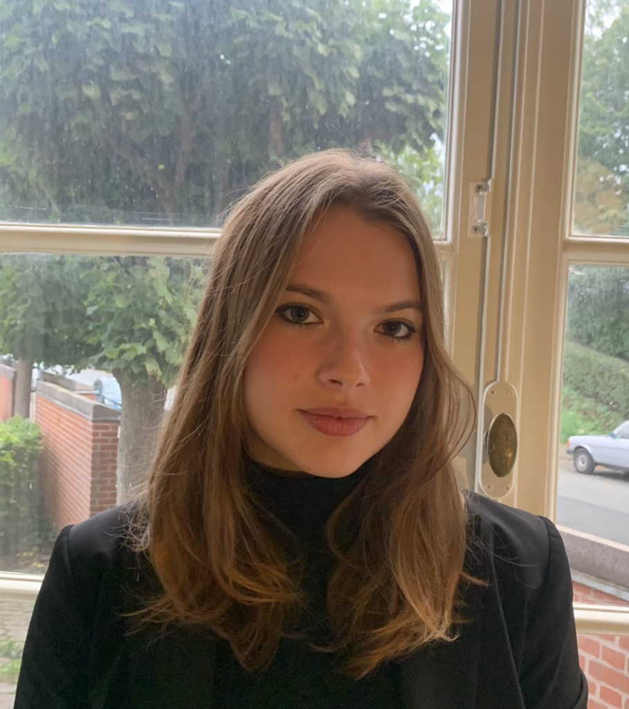 A young woman with light brown hair wearing a black top and blazer poses indoors in front of a window, embodying storytelling. Outside, trees, a brick wall, and a parked car are visible on a cloudy day.