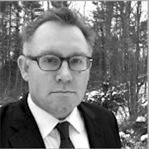 A man wearing glasses and a suit stands outdoors in a snowy, wooded area. The image is in black and white.