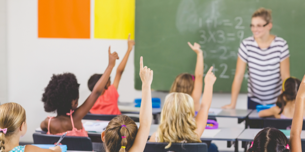 A group of young students sit at desks in a classroom, raising their hands to answer a question. Despite issues like absenteeism, a teacher stands at the front near a chalkboard with math problems written on it.