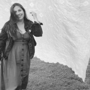 Kendra Carter stands on grass in a dress and denim jacket, smiling with one hand raised. A large, patterned sheet billows gently behind her. The black and white image captures a light and joyful moment.