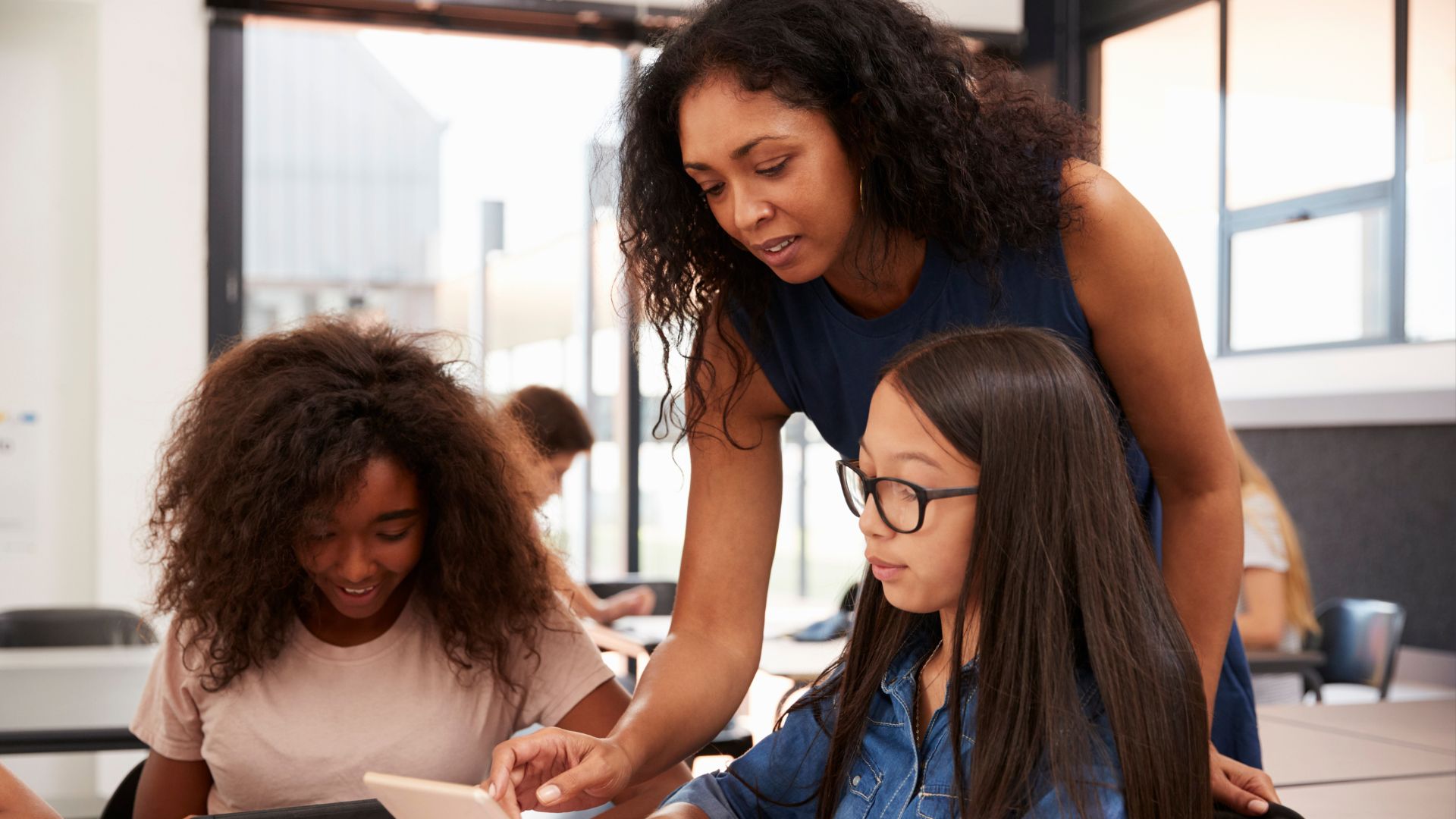 A teacher leans over to help two students with a tablet in a classroom, offering multi-tiered levels of support. One student points at the screen while the other looks on, as other students work independently in the background.