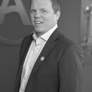 Black and white photo of a white man with short, sandy blond hair, in an ASU suit, standing in front of an A.