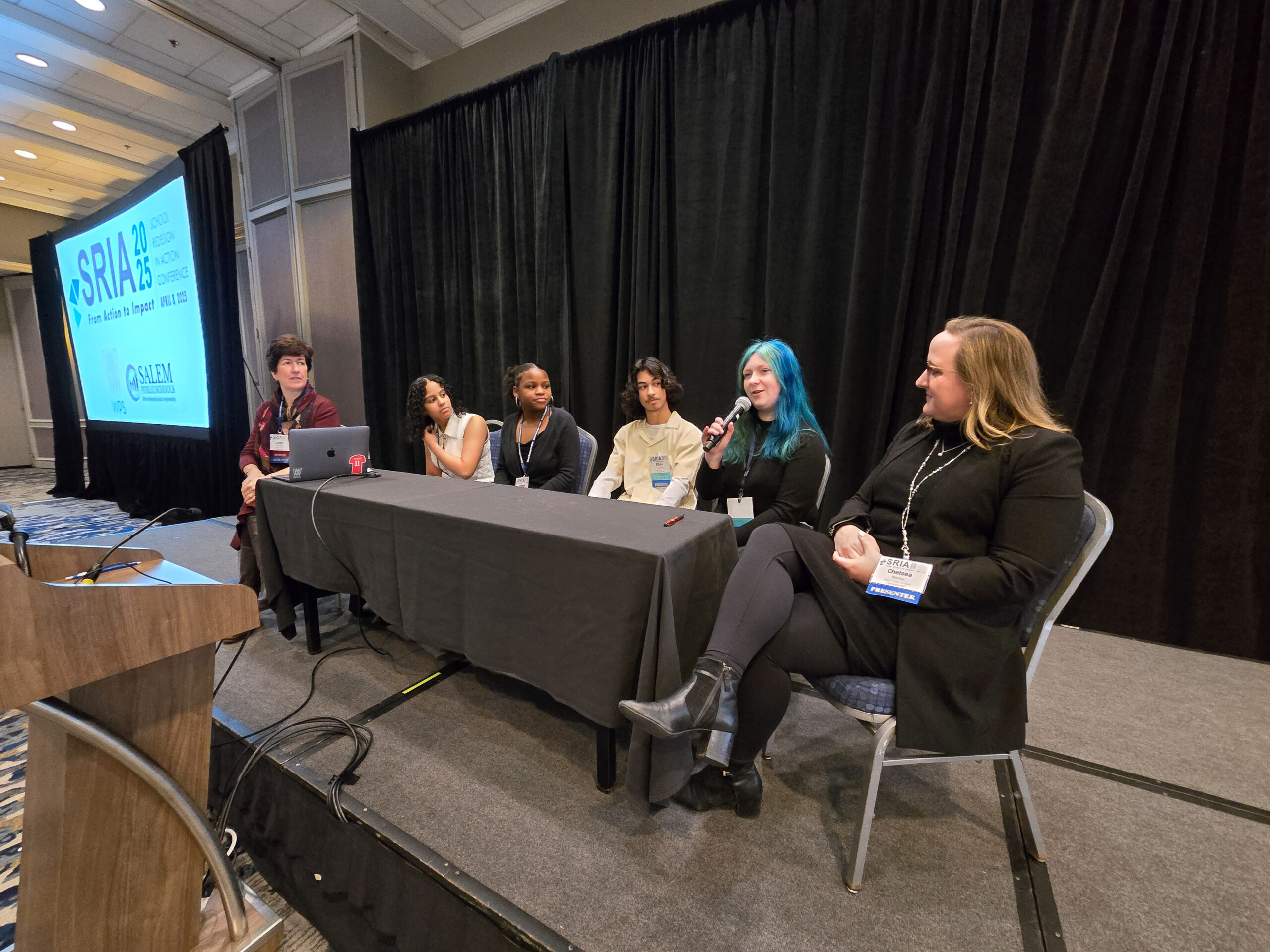 Six people sit at a long table on a stage in a conference room. One person with blue hair speaks into a microphone, while the projector screen displays “SSRIA 23” and SRIA presentation details.