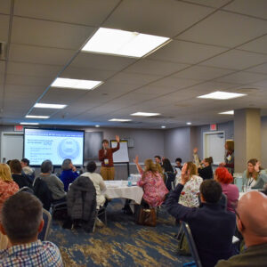 A group of people attend an SRIA workshop in a conference room, sitting at round tables. A presenter stands at the front, speaking and gesturing toward a projected slide as several attendees raise their hands.