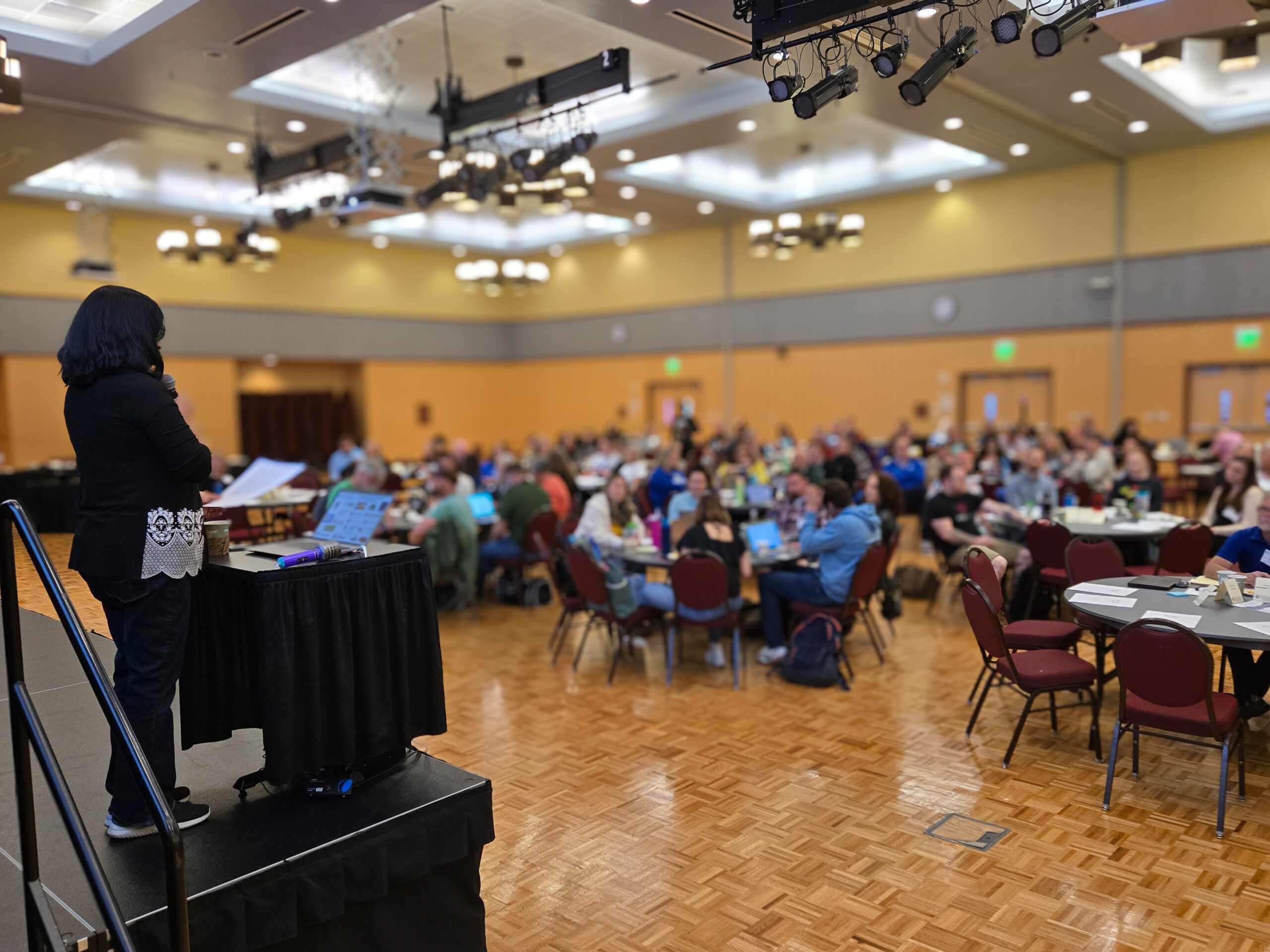 A person stands on a stage speaking to a large audience about Mastery Based Learning in a spacious, well-lit conference room. The crowd listens attentively, and presentation materials are visible on the podium.