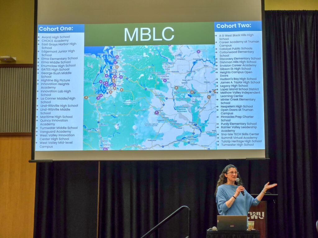 A presenter stands next to a projector screen displaying a map and two lists of school names under headings “Cohort One” and “Cohort Two,” with “MBLC” (Mastery Based Learning Collaborative) in large letters at the top.