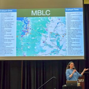 A presenter stands next to a projector screen displaying a map and two lists of school names under headings “Cohort One” and “Cohort Two,” with “MBLC” (Mastery Based Learning Collaborative) in large letters at the top.