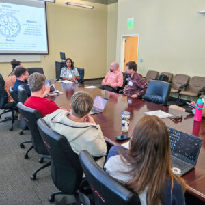 A group of people sit around a conference table in a meeting room, listening to a presenter discuss Mastery Based Learning. A presentation slide is displayed on the screen while some attendees use laptops and take notes.