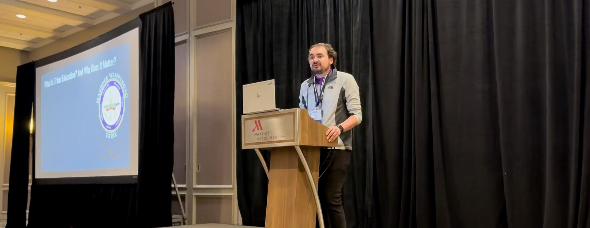 Author Brad Lopes stands at a podium, with a laptop in front of him and a screen to his right that reads "What is tribal education and why does it matter?"